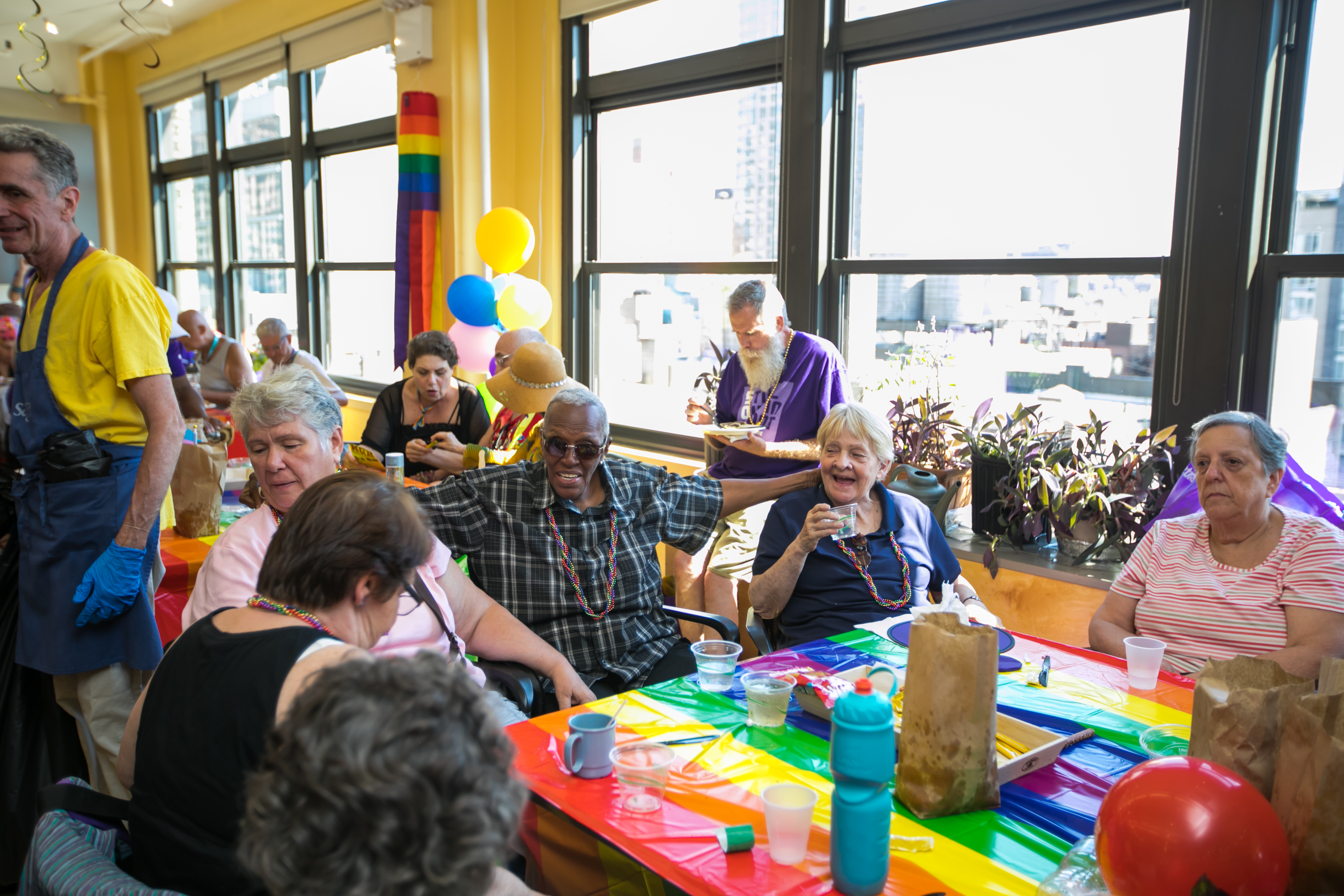 Group of LGBTQ+ older adults seated at a table covered in a rainbow table cloth at a party